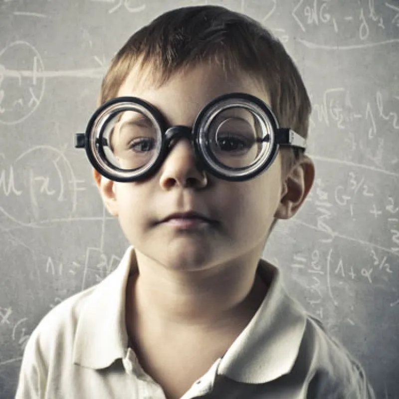 A young boy in thick round glasses in front of a chalkboard of formulas